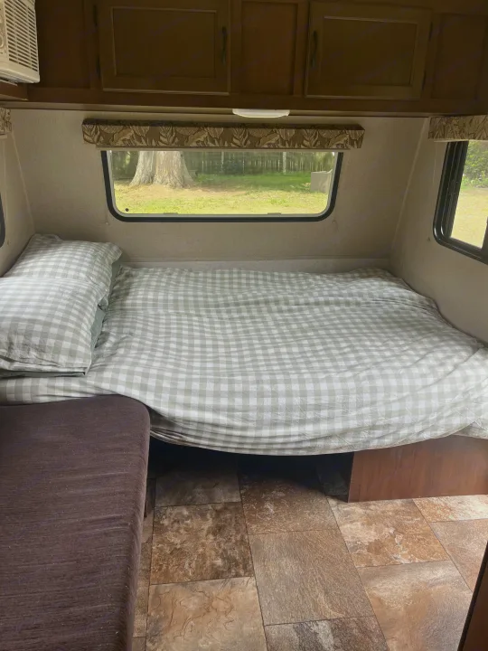 RV rental interior featuring a bed with sage green checkered bedding, nature views from windows, and wood cabinets.. Jayco Jay Flight 2015