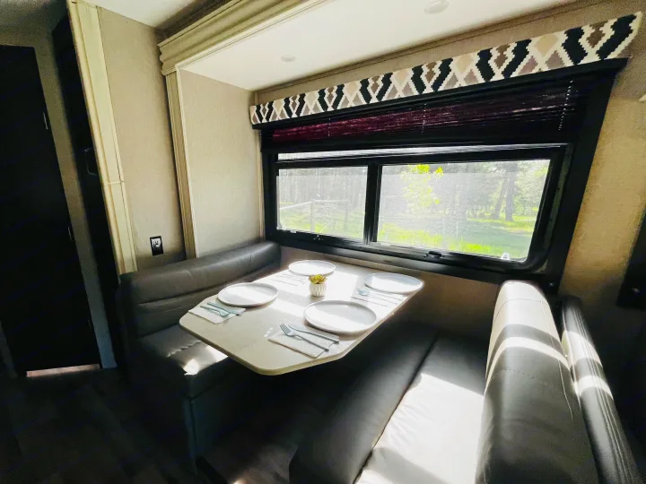 Bright rv rental dinette with grey upholstered benches, a set table for four, and a scenic forest view through the window.. Entegra Coach Odyssey 2021