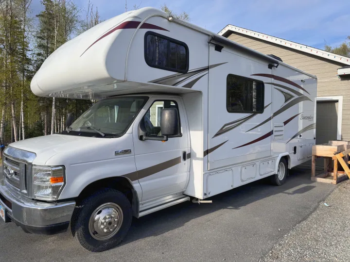 A white Class C RV rental, a Sunseeker by Forest River, parked on asphalt with trees & a house in the background.. Forest River Sunseeker 2018