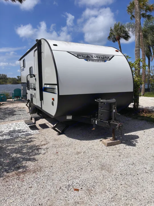 A white and black rv rental travel trailer is set up by a sunny waterfront, complete with palm trees and a view of the water.. Forest River Salem FSX PLATINUM 2020