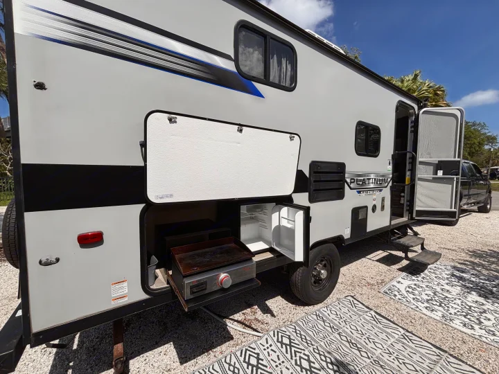 Grey RV rental with an open outdoor kitchen, revealing a griddle and mini-fridge. Decorative rugs lie on the gravel ground.. Forest River Salem FSX PLATINUM 2020