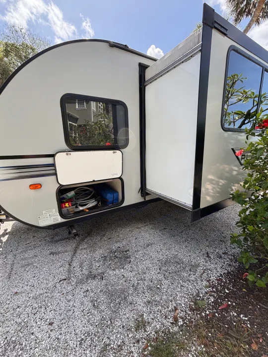An RV rental, light-grey and parked on gravel. Its slide-out is extended, and a storage hatch shows camping gear inside.. Forest River Salem FSX PLATINUM 2020