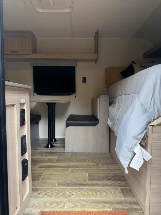 Modern RV rental interior with a dinette, TV, and comfortable bed, featuring light wood finishes and tile flooring.. Heartland RVs Pioneer 2025