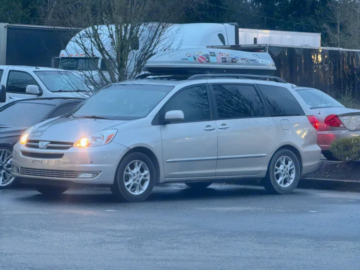 Silver minivan with a roof cargo box and bright headlights, parked near commercial trailers, suggesting an RV rental area.. Toyota Sienna Limited XLE Camper Van 2004