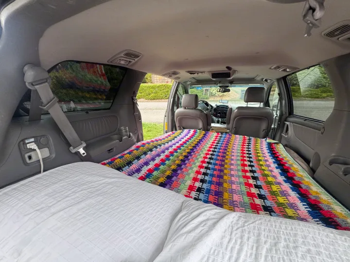 View from rear of a minivan converted for car camping or an RV rental. A comfortable bed with white bedding and a colorful crochet blanket fills the back.. Toyota Sienna Limited XLE Camper Van 2004