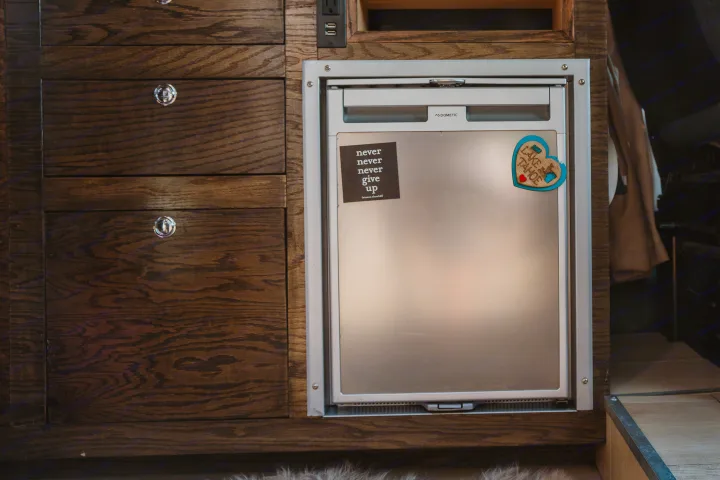 An RV rental's Dometic compact fridge & dark wood cabinets. Features a "Never Give Up" sticker and Lake Tahoe magnet.. Dodge 1500 2020