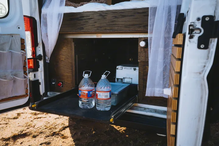 RV rental: rear of camper van, bed above storage. Pull-out tray holds two water jugs & a portable toilet. Sheer curtains visible.. Dodge 1500 2020