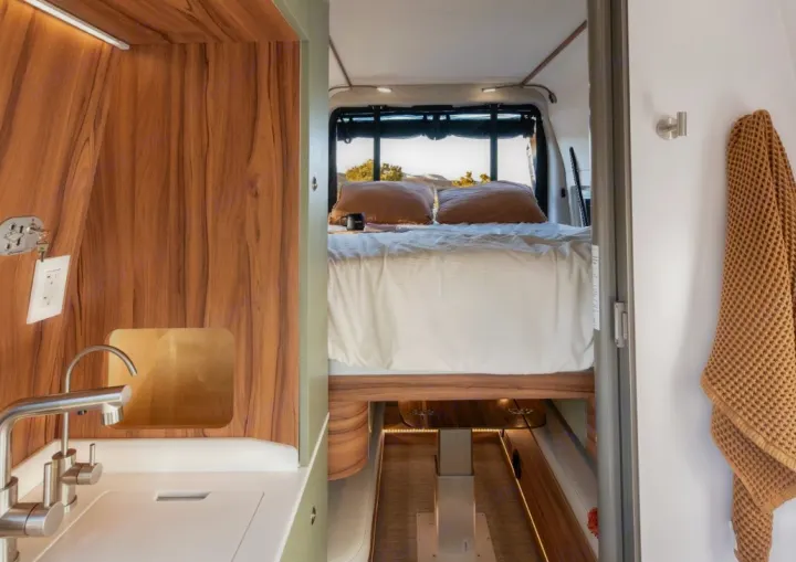This rv rental interior features a cozy bed with a scenic view, a functional sink, and stylish wood accents, ideal for travel.. Noovo Plus LB 2025
