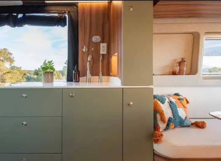 This rv rental features a modern kitchen with sage green cabinets, a sink, and a cozy lounge area with a scenic view.. Noovo Plus LB 2025