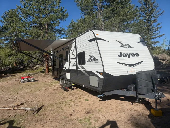 A modern Jayco travel trailer RV rental, with an extended awning, parked in a sunny pine forest with a red dirt bike.. Jayco Jay Flight SLX 2022