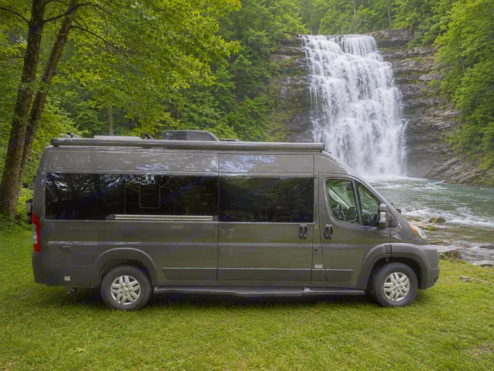 Grey rv rental camper van parked on grass, beside a river & grand waterfall. Lush green trees surround the stunning nature scene.. Thor Sequence 2022