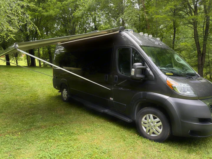 Dark gray campervan with an extended awning parked on green grass amidst trees, perfect for an RV rental getaway.. Thor Sequence 2022