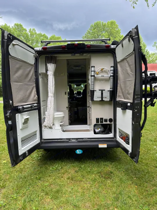 Open rear doors of an RV rental, revealing a compact interior with a toilet and organized storage for camping gear.. Thor Sequence 2022