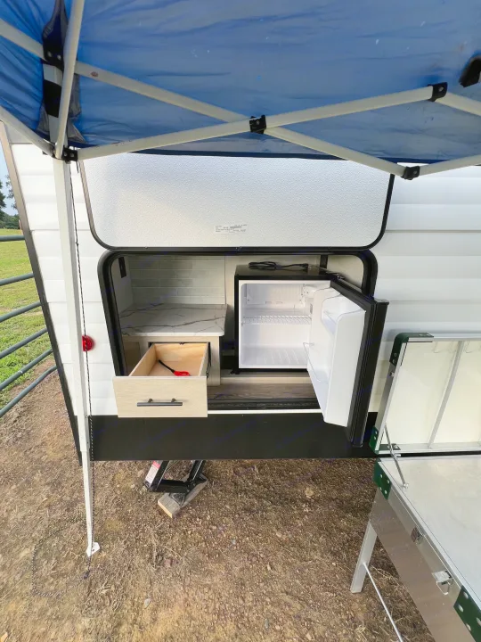 An RV rental's open outdoor kitchen with a mini-fridge, marble-look counter, and open drawer under a blue canopy.. Forest River Shasta I-5 2026