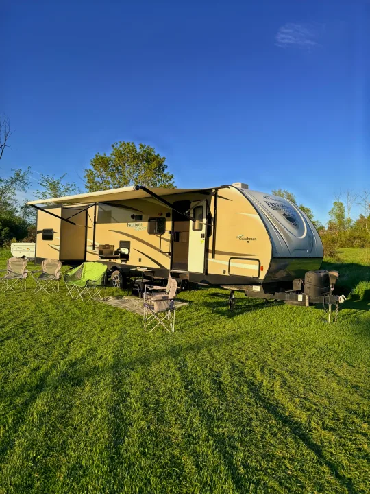 Relaxing with an RV rental! This tan travel trailer is set up in a sunny, grassy field with chairs for outdoor camping.. Coachmen Freedom Express 312BHDS Travel Trailer 2017