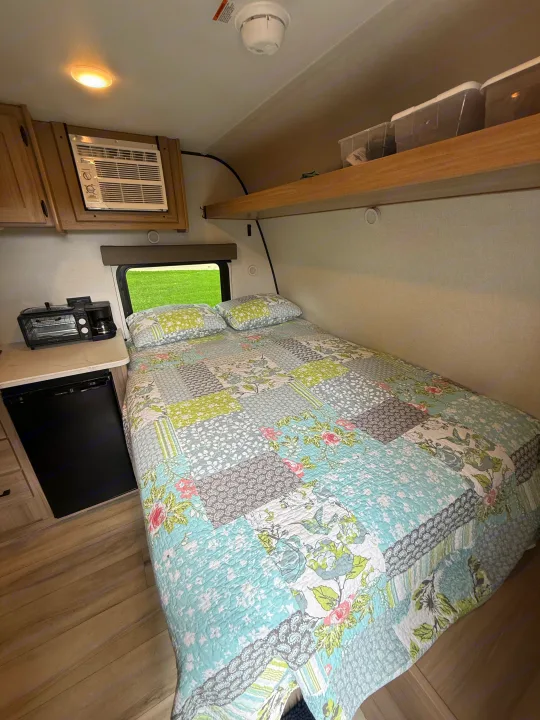Cozy rv rental: a bedroom and kitchenette area with a colorful quilt, compact AC, mini-fridge, and toaster oven combo.. Keystone RV Coleman 13B 2026