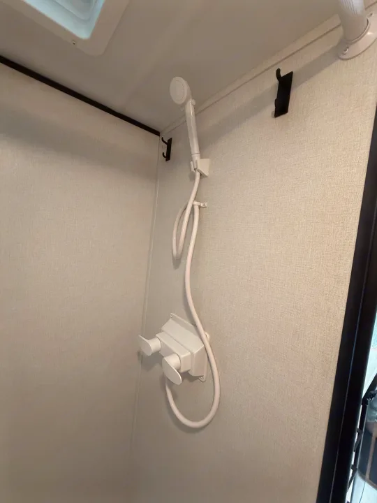 Interior of an RV rental shower featuring a white handheld showerhead, hose, and dual controls on a light textured wall with hooks.. Keystone RV Coleman 13B 2026