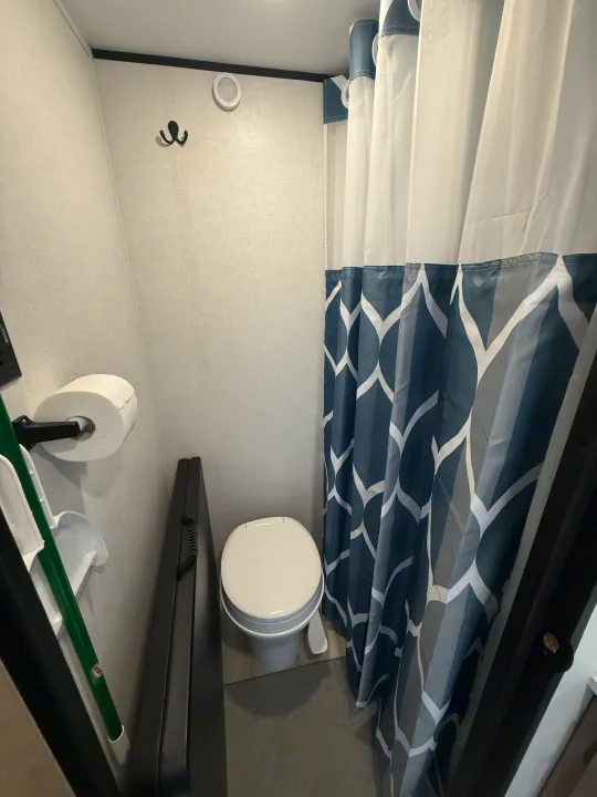 Compact RV rental bathroom with a toilet, shower curtain featuring blue and white geometric patterns, wall hook, and toilet paper.. Keystone RV Coleman 13B 2026