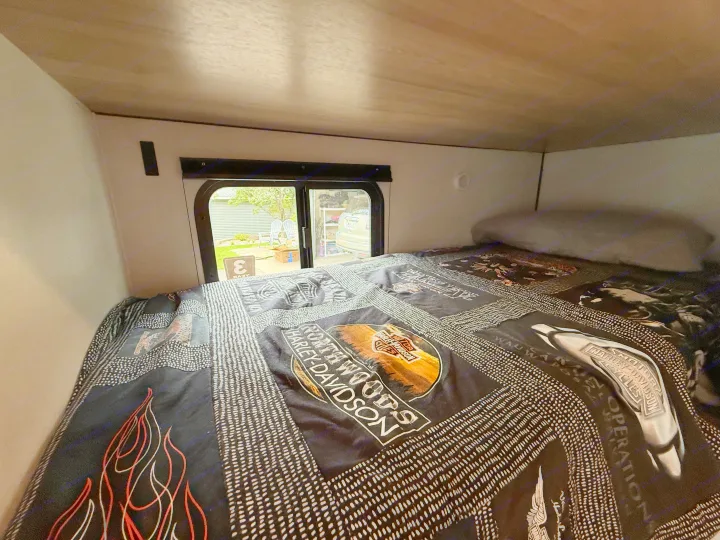 Cozy RV rental bed with a Harley-Davidson quilt, offering a window view of a residential area.. Keystone RV Coleman 13B 2026