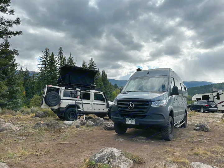A white SUV with a rooftop tent & a grey camper van (likely an RV rental) parked at a rugged mountain campsite under cloudy skies.. Mercedes-Benz Sprinter 2020