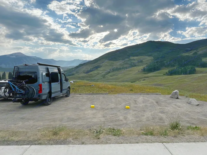 Grey RV rental campervan with bikes parked at a scenic mountain overlook under dramatic, cloud-filled skies over vast green hills.. Mercedes-Benz Sprinter 2020