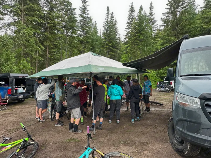 Muddy mountain bikers gather under tents and an RV rental awning in a forest. Bikes and vehicles suggest an outdoor adventure.. Mercedes-Benz Sprinter 2020