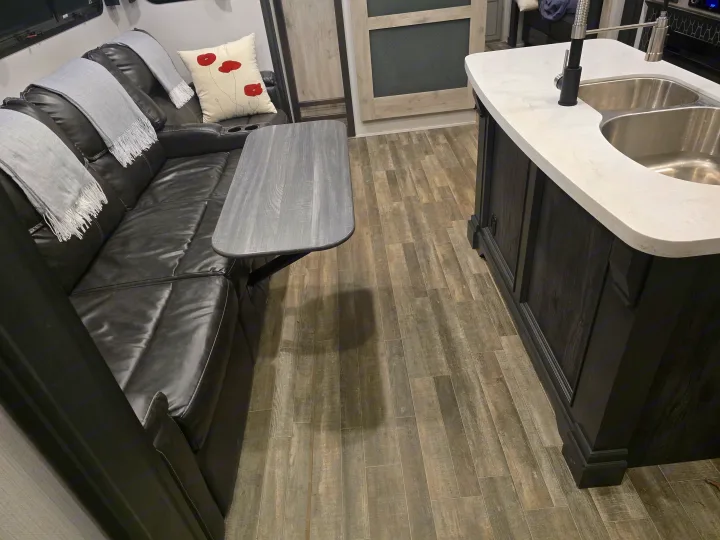 Inside an RV rental: a black leather sofa with a pull-out table, and a kitchen island with sink, on wood-look floors.. Keystone RV Alpine Avalanche 2024