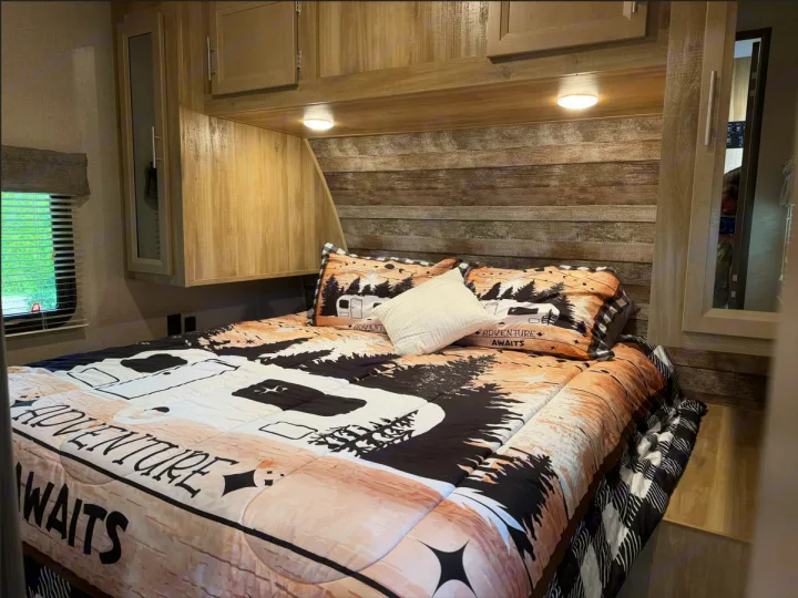 This cozy RV rental bedroom features rustic decor, a bed with an "Adventure Awaits" comforter, and ample overhead storage.. Coachmen Catalina Legacy 2022