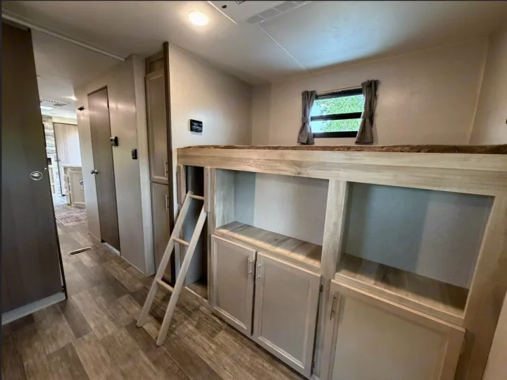 An RV rental features bunk beds, a ladder for access, and built-in storage cabinets below. A window and hallway are also visible.. Coachmen Catalina Legacy 2022