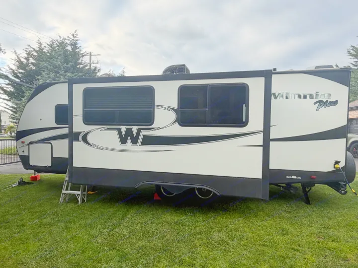 White Winnebago Minnie Plus travel trailer (RV rental) with its slide-out extended, parked on green grass under a cloudy sky.. Minnie Minnie Plus 2019