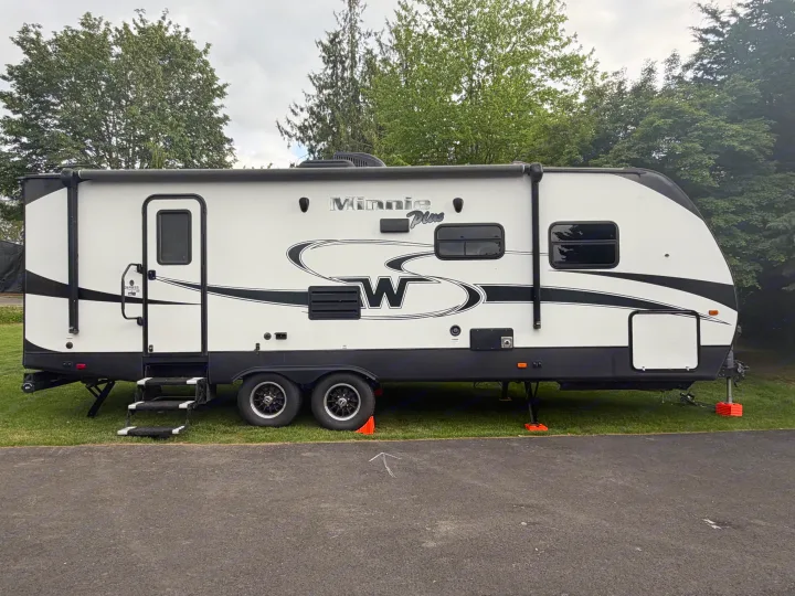 A white and black Minnie Plus travel trailer RV rental parked on green grass next to an asphalt road, surrounded by trees.. Minnie Minnie Plus 2019