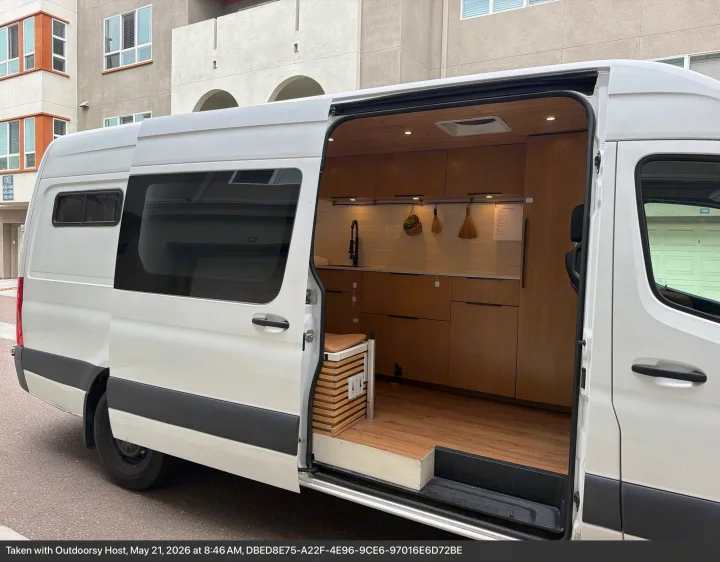Open side door of a white rv rental van reveals a sleek wooden interior with a kitchen, sink, and cabinets.. Mercedes-Benz Sprinter 2020