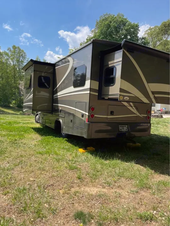 Large motorhome (RV rental) parked on grass with extended slide-outs, leveling blocks, trees & blue sky.. Isata 3 2017