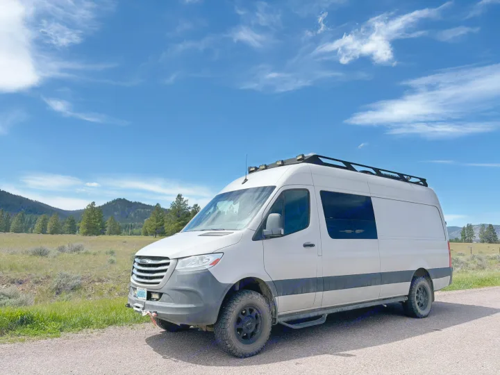 A white off-road camper van is parked on a gravel road with mountains and blue sky. Perfect for an rv rental adventure!. Mercedes Sprinter 2019