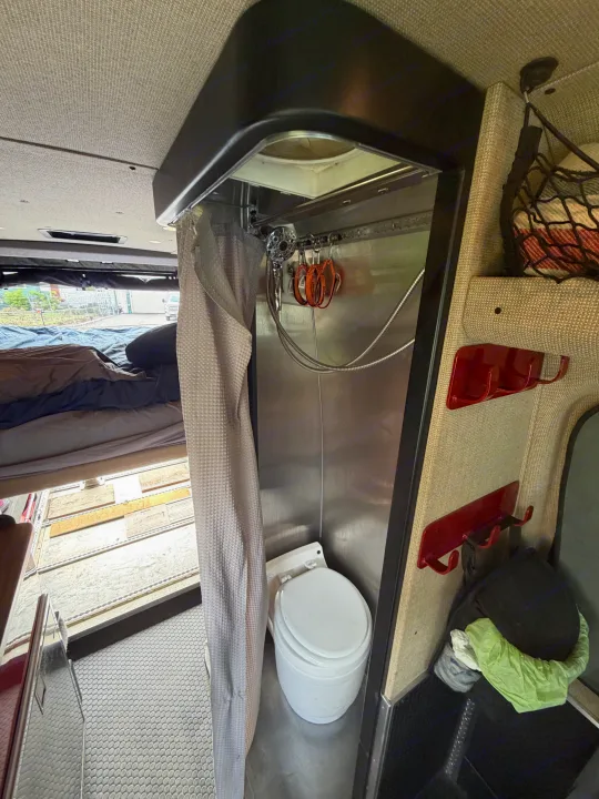 The interior of an RV rental showing a compact bathroom with a toilet and shower, next to an elevated bed. Textured walls and smart storage are visible.. Mercedes Sprinter 2019