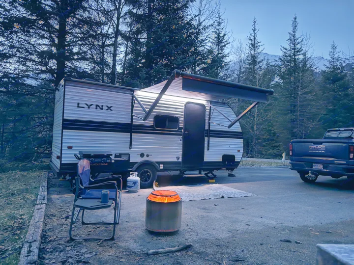 Evening at a serene RV rental campsite. A lit travel trailer, glowing fire pit, camping chair, and distant snow-capped mountains.. Heartland RVs Prowler Lynx 2024