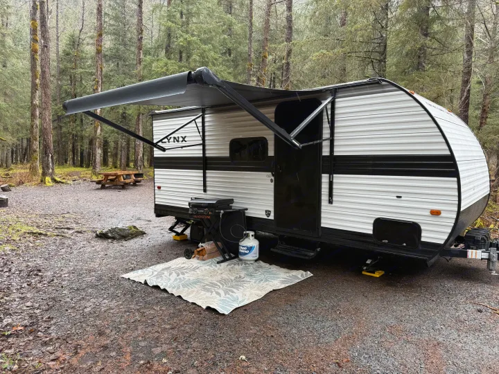 This RV rental, a white and black travel trailer, is set up at a forest campground with its awning extended, featuring a grill and outdoor rug.. Heartland RVs Prowler Lynx 2024