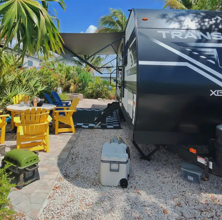 Scenic rv rental spot with an extended awning, outdoor table, chairs, cooler, and lush tropical foliage.. Grand Design Transcend Xplor 2026