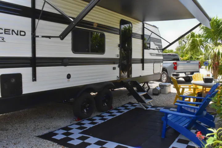 This RV rental offers a cozy outdoor setup with an extended awning, steps, and chairs on a checkered mat at a sunny campsite.. Grand Design Transcend Xplor 2026