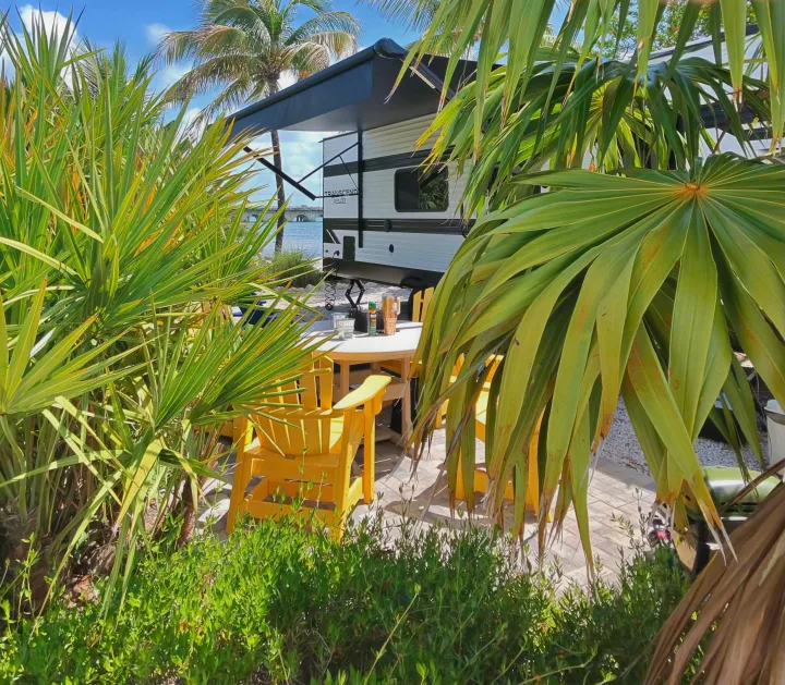 Scenic rv rental site: a white travel trailer under an awning, surrounded by vibrant palm trees & yellow chairs near the water.. Grand Design Transcend Xplor 2026
