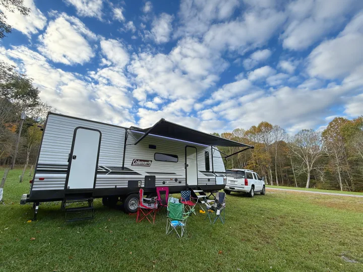 RV rental camping trip: A Coleman travel trailer with an extended awning, chairs, and SUV under a cloudy sky and autumn trees.. Dutchmen Coleman Lantern LT 2024
