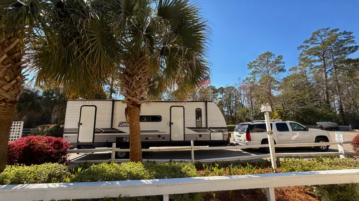 White Coleman RV rental trailer and SUV parked. Sunny day with palm and pine trees. An American flag flies nearby.. Dutchmen Coleman Lantern LT 2024