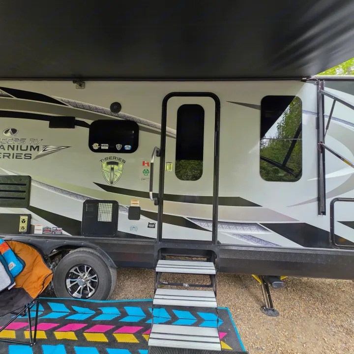 Enjoying an RV rental: a detailed view of the Outdoors RV trailer's side, showing its extended awning, entry, and campsite mat.. Outdoors RV 23DBS 2023