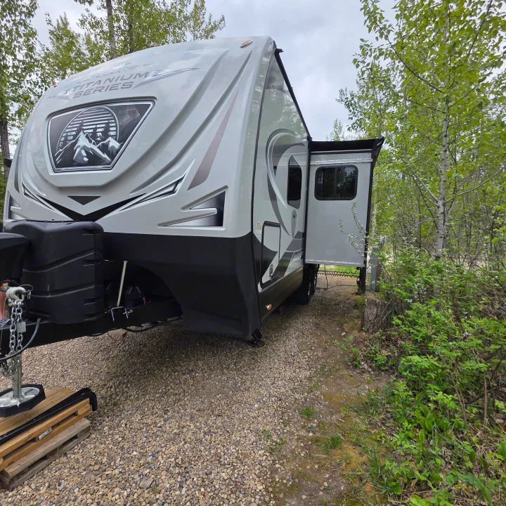 Grey Titanium Series travel trailer rv rental parked on gravel amidst green trees, ideal for a nature camping trip.. Outdoors RV 23DBS 2023