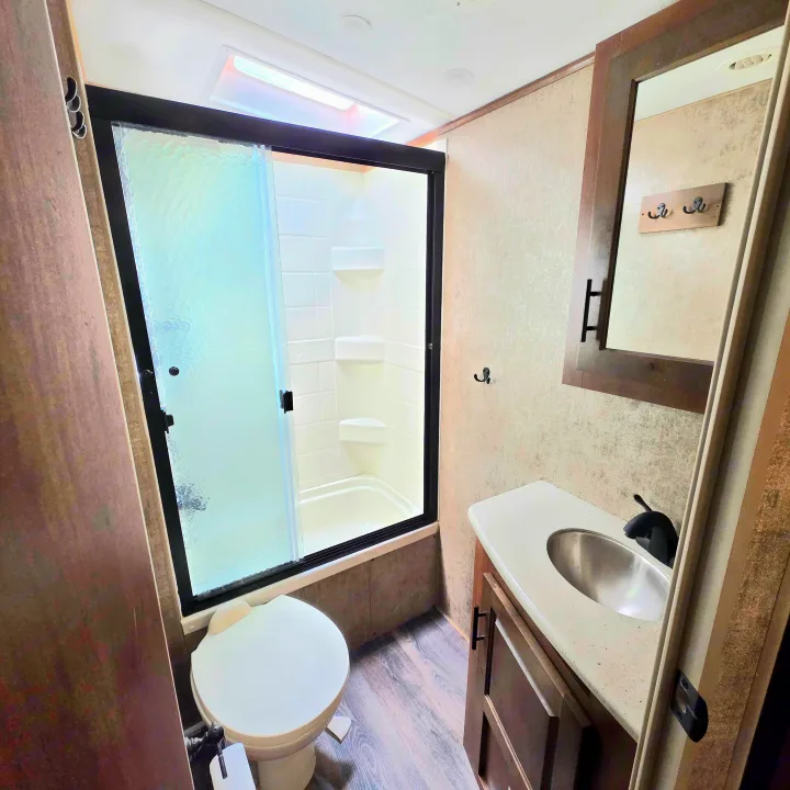 Clean RV rental bathroom features a glass-enclosed shower, toilet, and vanity with sink and mirror.. Outdoors RV 23DBS 2023