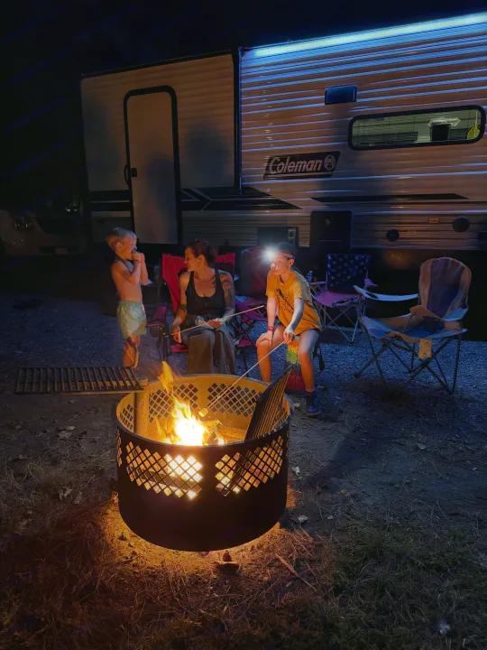 Family roasts marshmallows by a glowing campfire with their RV rental under blue awning lights. Cozy camping night.. Dutchmen Coleman Lantern LT 2024