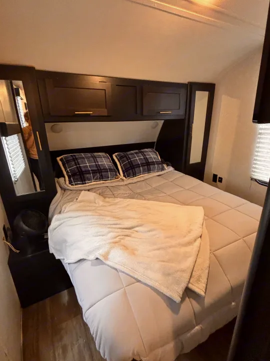 Cozy RV rental bedroom featuring a full bed with plaid pillows, overhead cabinets, and side mirrors, perfect for travel.. Forest River Cherokee Grey Wolf 2021