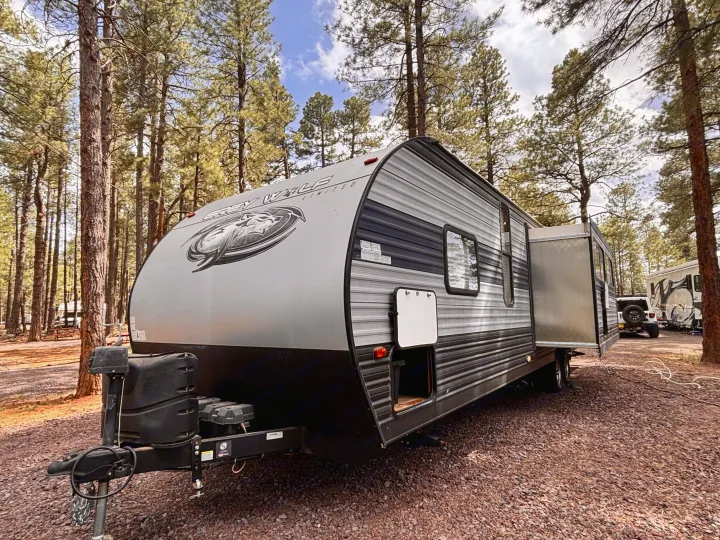 Grey Wolf travel trailer RV rental parked in a peaceful pine forest campsite, surrounded by tall trees and gravel ground.. Forest River Cherokee Grey Wolf 2021