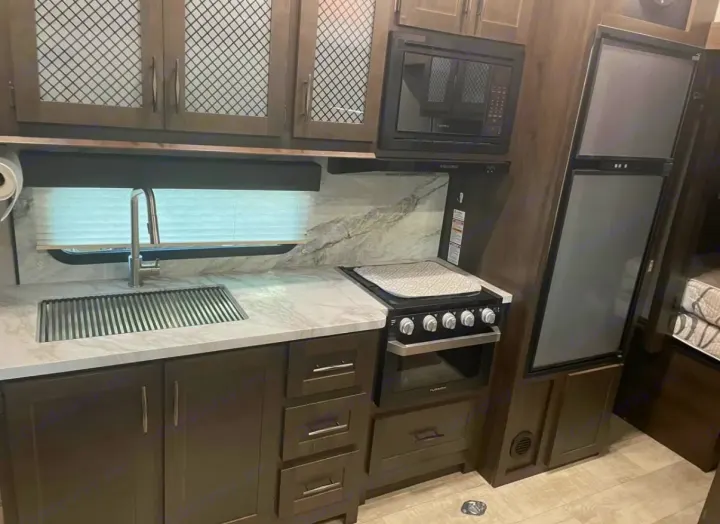 Modern RV rental kitchen with sleek sink, marble-look counter, microwave, oven/stove, and fridge.. Grand Design Momentum 2021