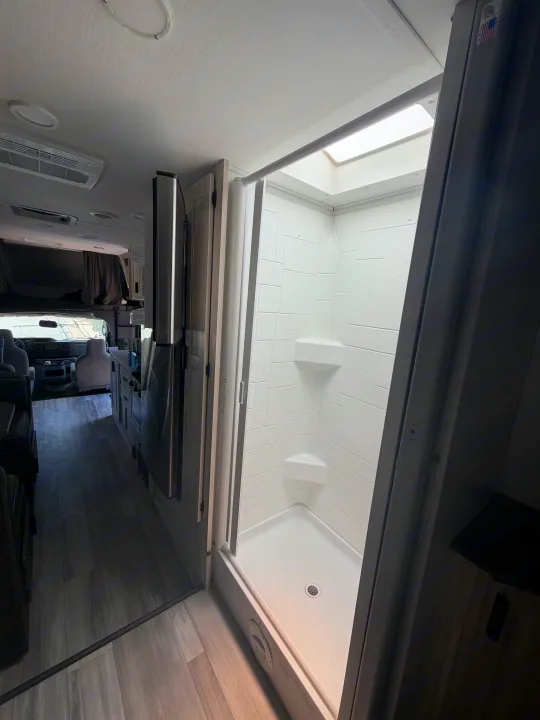 Step into this RV rental: bright, tiled shower in the foreground, with a galley kitchen, fridge, and cockpit receding into the back.. Forest River Sunseeker LE 2024